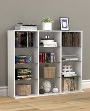 Weißes, modernes Regal mit neun Fächern und Glasböden, dekoriert mit Büchern, Pflanze, Bilderrahmen und Dekoobjekten; frontale Ansicht auf grauer Wand und Holzboden.