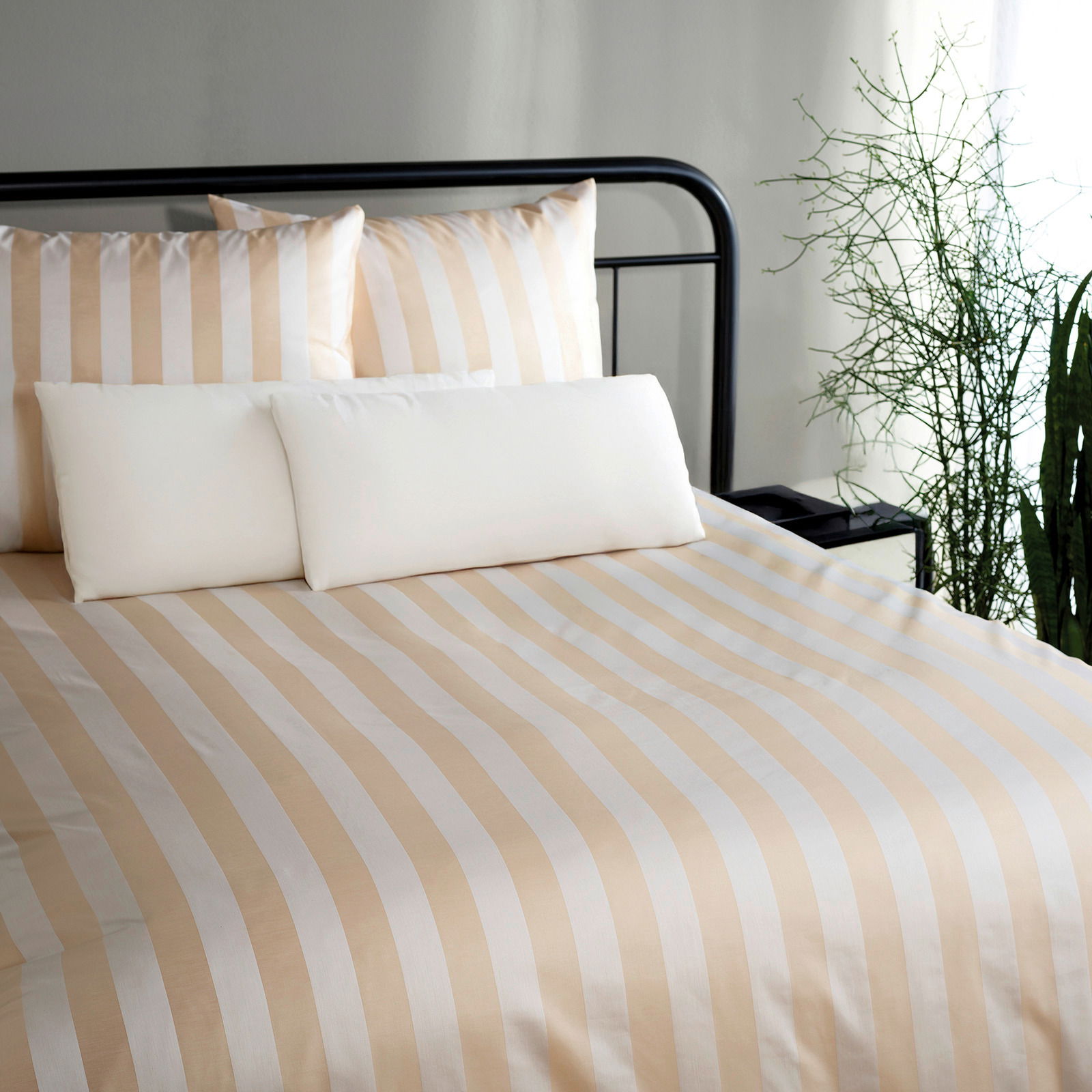 Bettwäschen Set mit beige-weißen Streifen, frontal fotografiert, auf einem schwarzen Metallbettgestell, umgeben von Pflanzen.