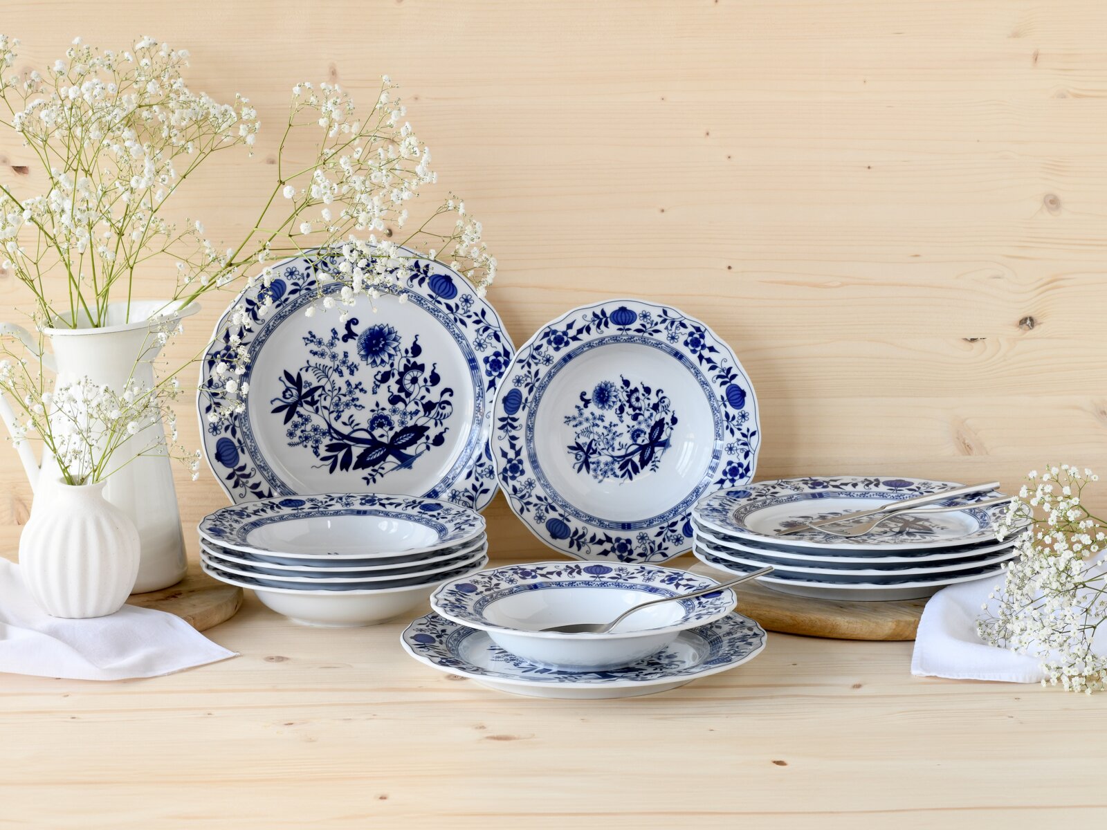 Blau-weißes Porzellan-Geschirrset mit floralem Dekor (Teller und Schalen) auf hellem Holztisch, dekoriert mit weißen Blumen und Besteck, aufgenommen in leicht erhöhter Frontalansicht.