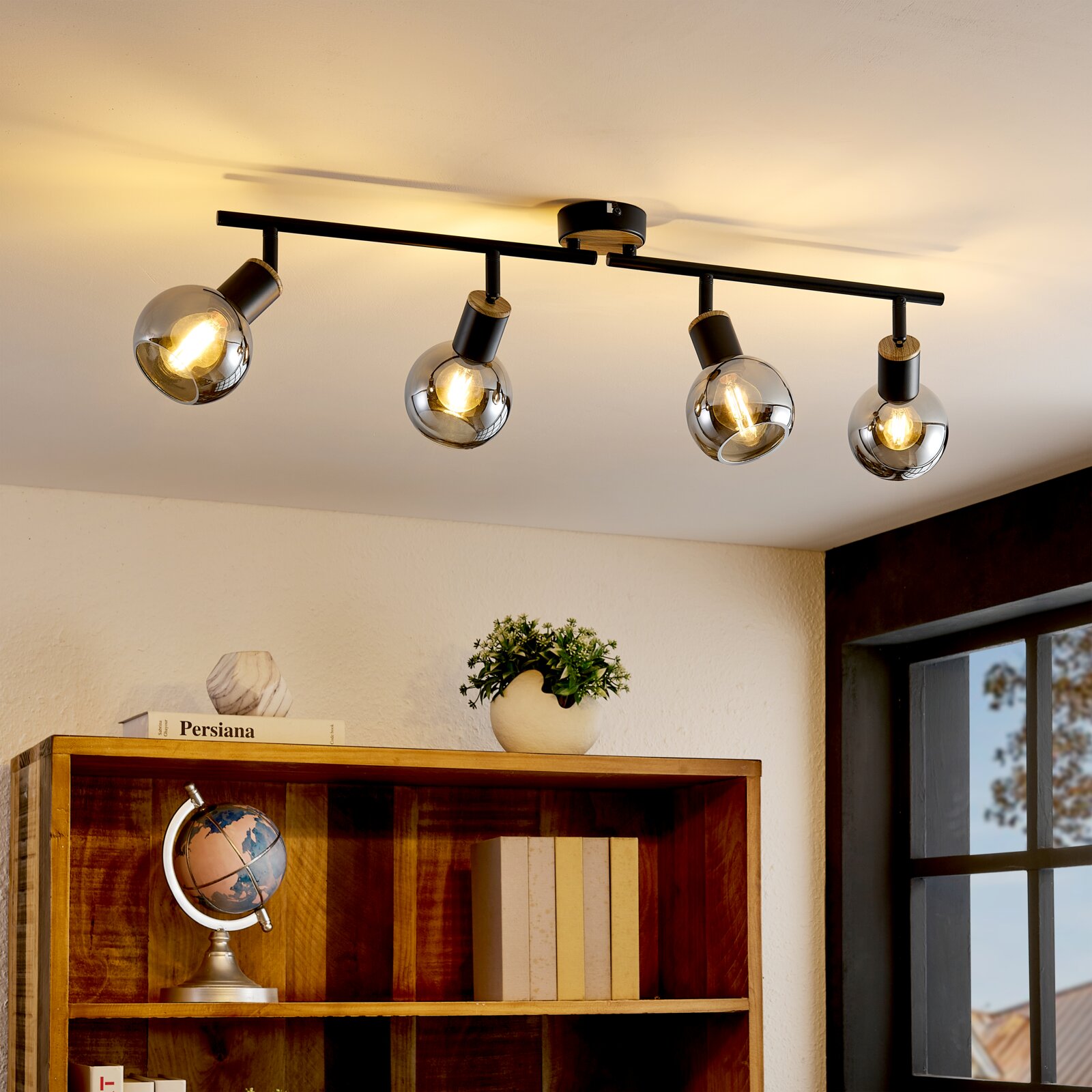 Moderne Deckenleuchte/Spotleiste mit vier schwenkbaren Strahlern, schwarzer Metallstange mit Holzdetails und rauchgrauen Glaskugelschirmen, warm leuchtend, aus leicht schräger Untersicht im Wohnraum fotografiert