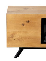 Nahaufnahme eines Holz-Kamins mit natürlicher Maserung und Astlöchern, schwarzem Metallfuß und teilweise sichtbarer Glasfront, Ansicht schräg von vorne