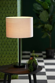 Moderner Lampenfuß mit rundem, beigem Lampenschirm auf einem Tisch, umgeben von grüner Dekoration. Perspektive: seitlich.