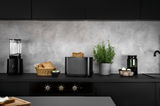 Schwarzer Toaster mit zwei Scheiben Toast, frontal auf einer modernen Küchenarbeitsplatte platziert, umgeben von einem Mixer, einem Brotkorb und einer Pflanze.