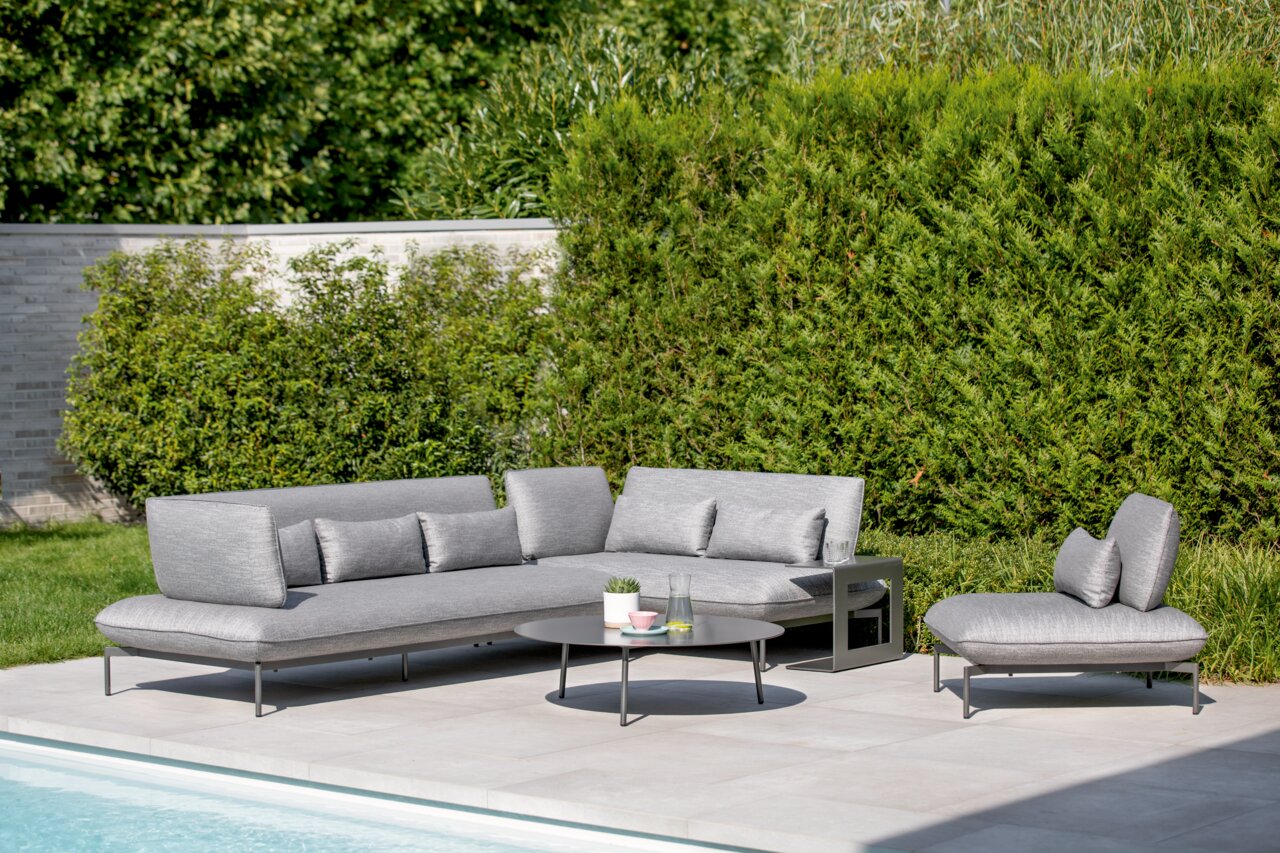 Gartenlounge-Set mit grauem Loungesofa, Loungesessel und rundem Beistelltisch, umgeben von grüner Hecke, aus der Perspektive eines Betrachters am Pool.