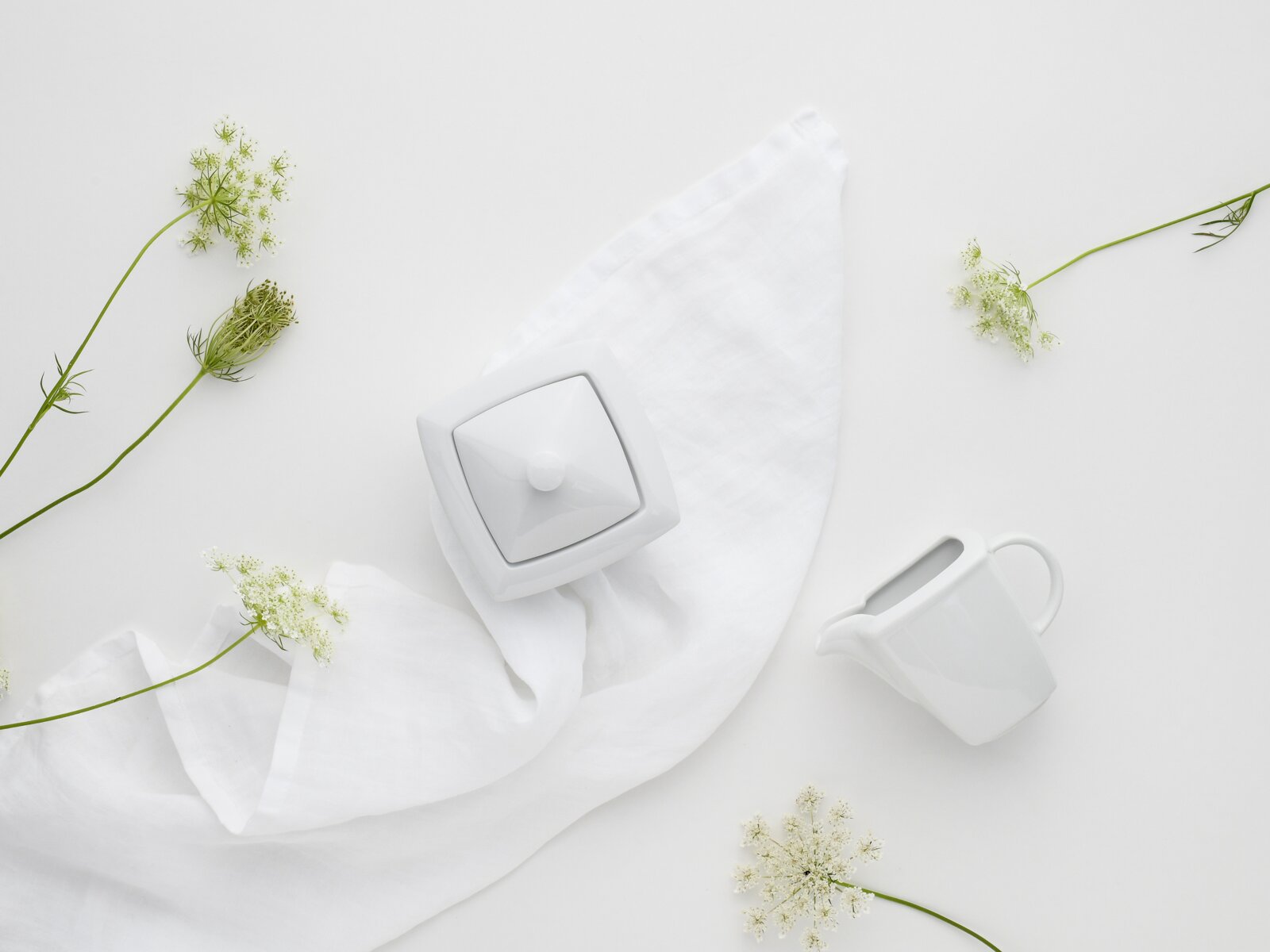 Minimalistisches Stillleben aus weißem Porzellan: quadratische Zuckerdose mit Deckel und kleines Milchkännchen auf weißem Stoff, umgeben von zarten weißen Blüten, Draufsicht (Flatlay)