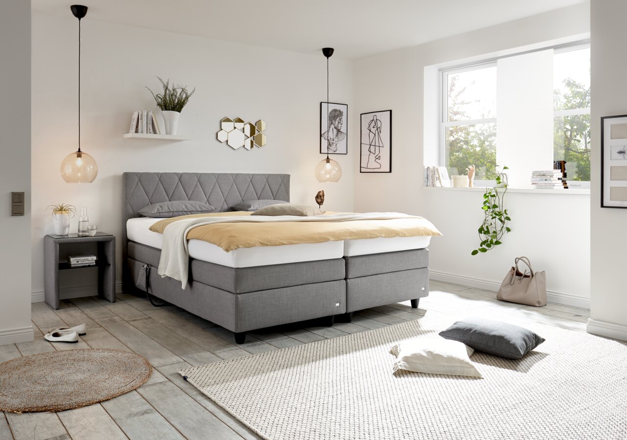 Modernes Boxspringbett in einem hellen Schlafzimmer, seitliche Perspektive, mit grauem Kopfteil und eleganter Dekoration.