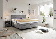 Modernes Boxspringbett in einem hellen Schlafzimmer, seitliche Perspektive, mit grauem Kopfteil und eleganter Dekoration.