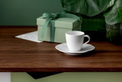 Weiße Kaffeetasse auf Untertasse auf einem Holztisch, daneben eine mintgrüne Geschenkbox mit Schleife und grüne Pflanze im Hintergrund, perspektivisch aus leicht seitlicher Ansicht auf Tischhöhe