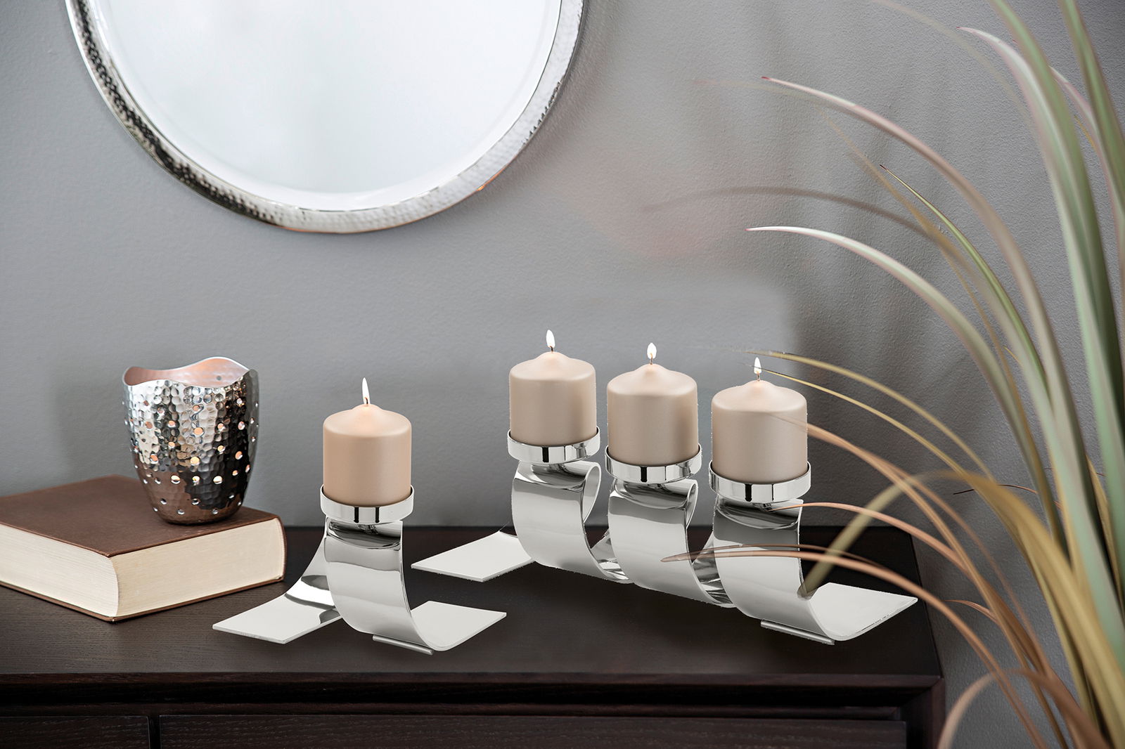 Moderner Kerzenhalter mit vier brennenden Kerzen, aus glänzendem Metall, auf einer dunklen Kommode vor einem Spiegel, seitliche Perspektive.