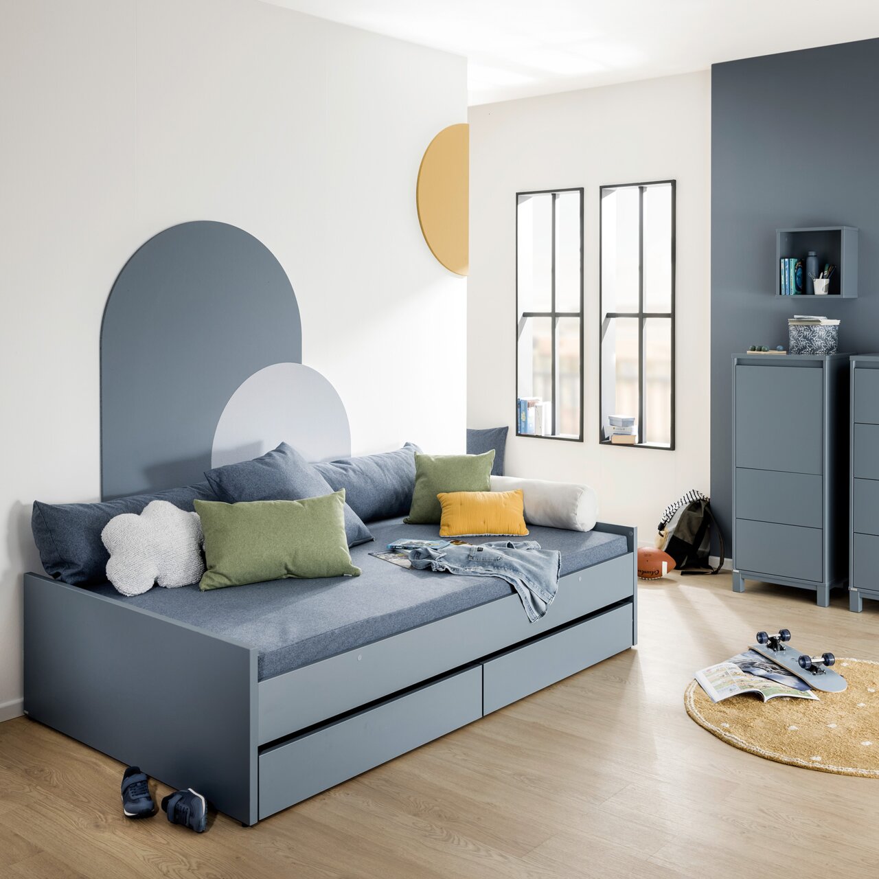 Modernes Bodenbett in einem stilvollen Raum mit blauen und grünen Kissen, zwei Schubladen und einem dekorativen Wanddesign. Perspektive von vorne links.