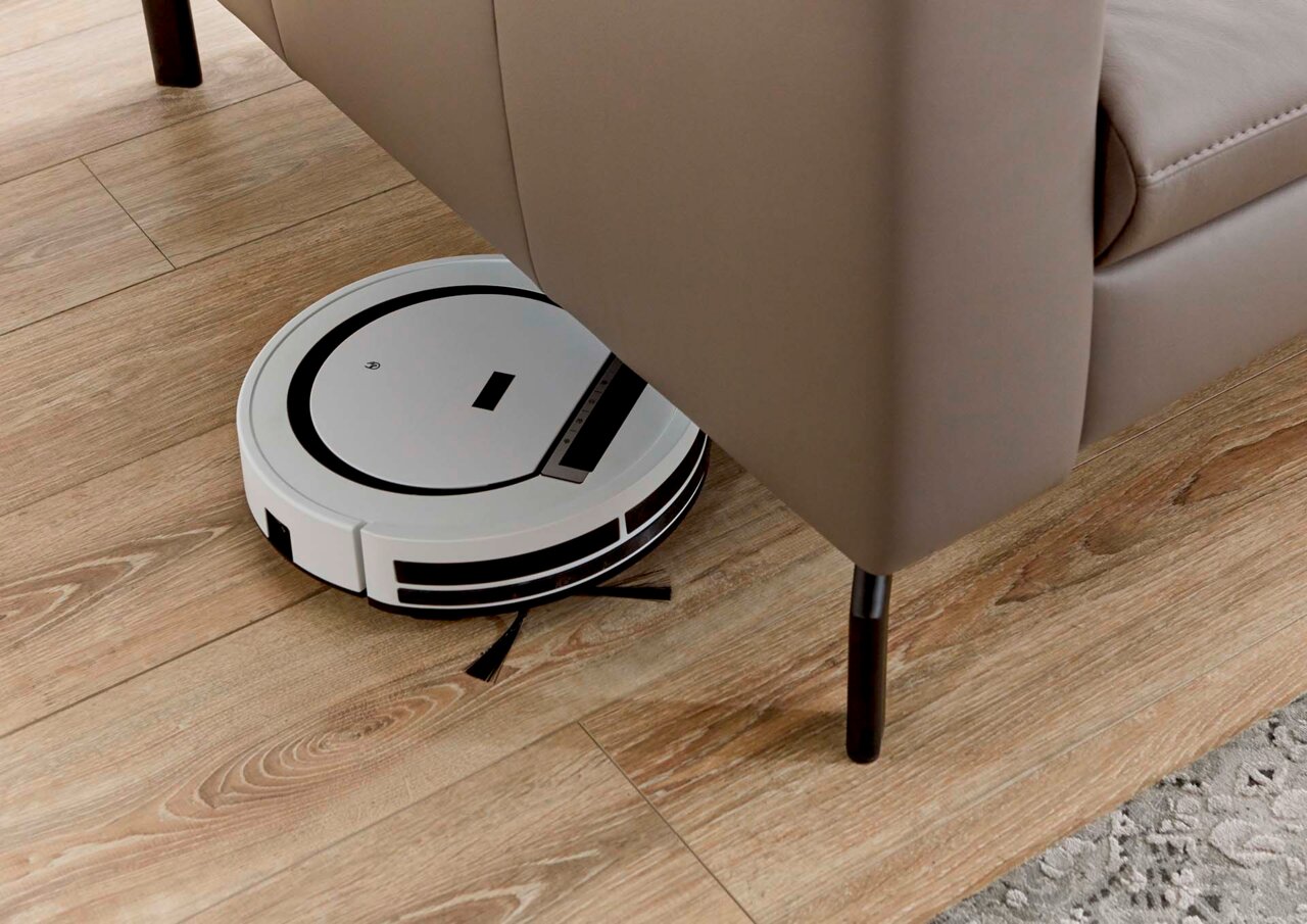 Een robotstofzuiger maakt de vloer onder een bank schoon, gezien vanuit de zijkant