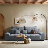 Großes, graues Big-Sofa mit mehreren Kissen, ein Hund sitzt darauf. Im Vordergrund ein runder Holztisch mit Dekoration. Perspektive: Frontalansicht.