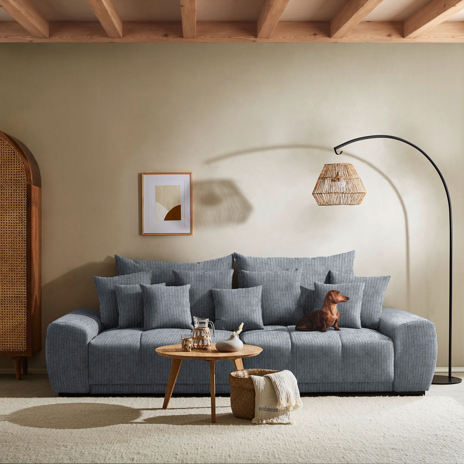 Großes, graues Big-Sofa mit mehreren Kissen, ein Hund sitzt darauf. Im Vordergrund ein runder Holztisch mit Dekoration. Perspektive: Frontalansicht.