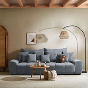 Großes, graues Big-Sofa mit mehreren Kissen, ein Hund sitzt darauf. Im Vordergrund ein runder Holztisch mit Dekoration. Perspektive: Frontalansicht.