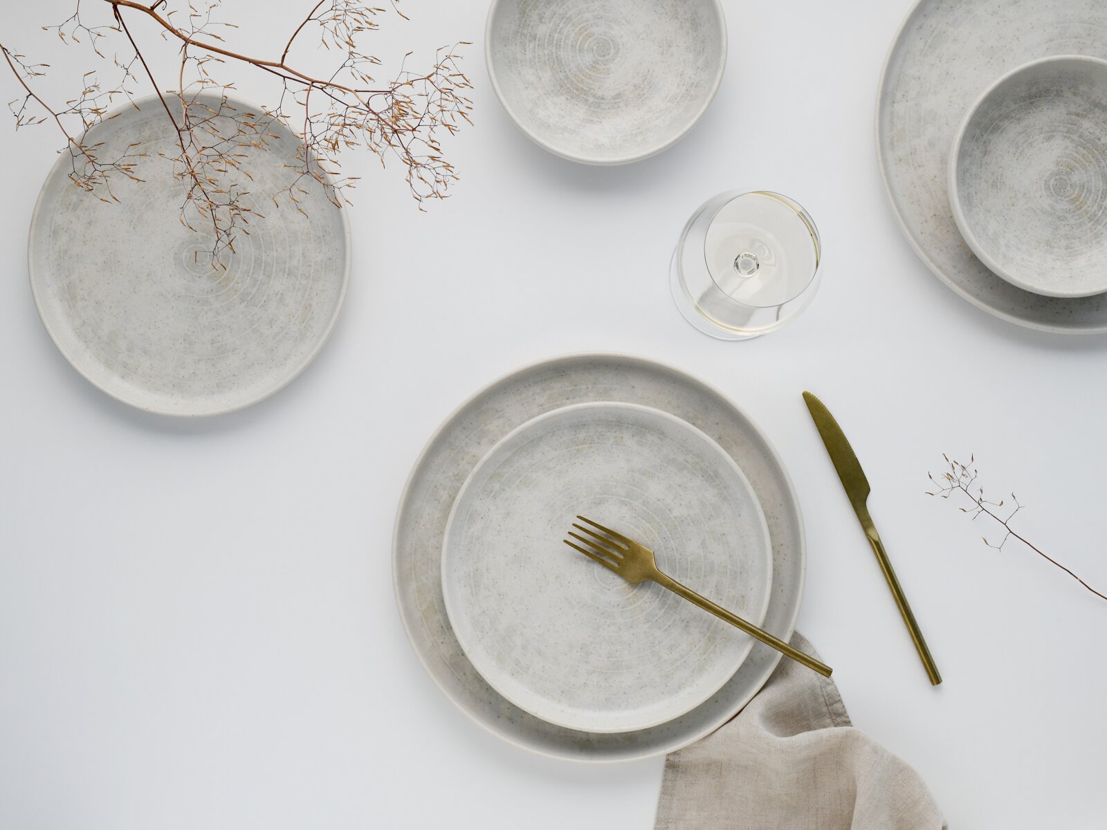 Minimalistisches Keramikgeschirr-Set in gesprenkeltem Hellgrau mit großen und kleinen Tellern sowie Schale, dazu goldfarbenes Messer und Gabel, Weinglas und Leinenserviette auf weißem Hintergrund, fotografiert in Draufsicht (Top-Down).
