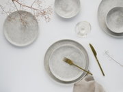 Minimalistisches Keramikgeschirr-Set in gesprenkeltem Hellgrau mit großen und kleinen Tellern sowie Schale, dazu goldfarbenes Messer und Gabel, Weinglas und Leinenserviette auf weißem Hintergrund, fotografiert in Draufsicht (Top-Down).