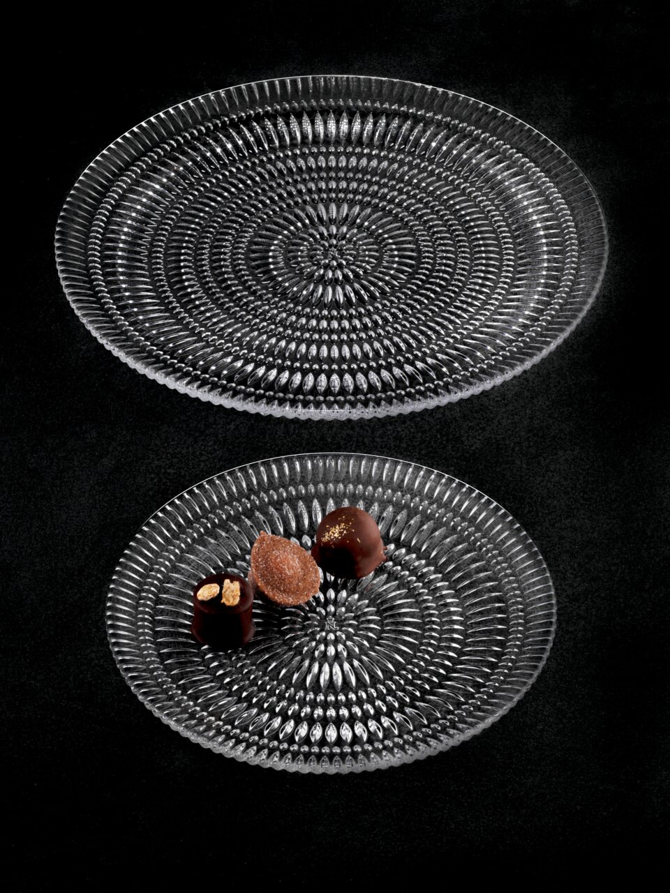 Zwei elegante, durchsichtige Glasteller mit strukturiertem Muster, von oben fotografiert. Auf dem kleineren Teller liegen drei Pralinen.
