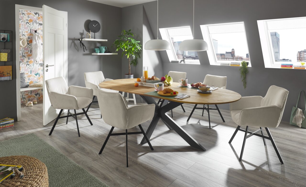 Moderner Esstisch mit ovaler Holzplatte und schwarzen Metallbeinen, umgeben von sechs beigen Stühlen in einem hellen Esszimmer mit Dachfenstern, seitliche Perspektive.