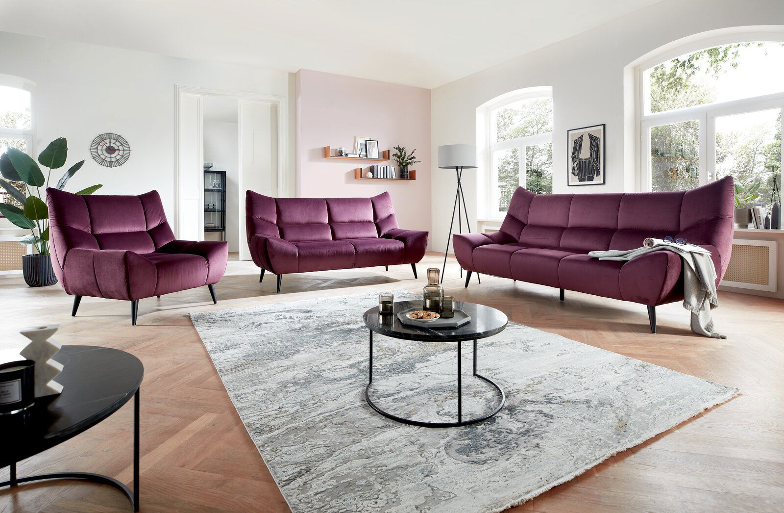 Wohnzimmer mit moderner Polstergarnitur in Lila, bestehend aus einem 3er-Sofa, einem 2er-Sofa und einem Sessel, aus der Perspektive von vorne betrachtet.