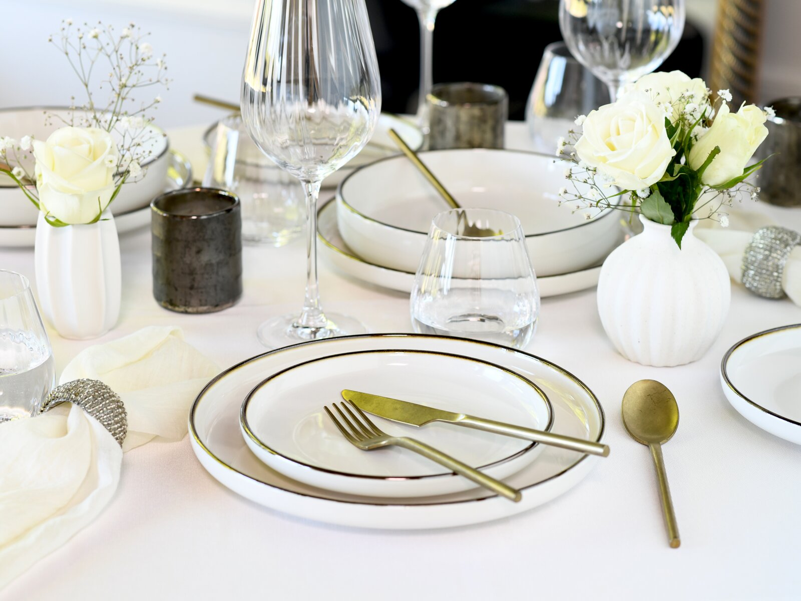 Elegant gedeckter Tisch mit weißem Geschirr mit feinem Goldrand, goldfarbenem Besteck, Wein- und Wassergläsern sowie weißen Rosen in Vasen, aufgenommen in leichter Schrägansicht von oben.