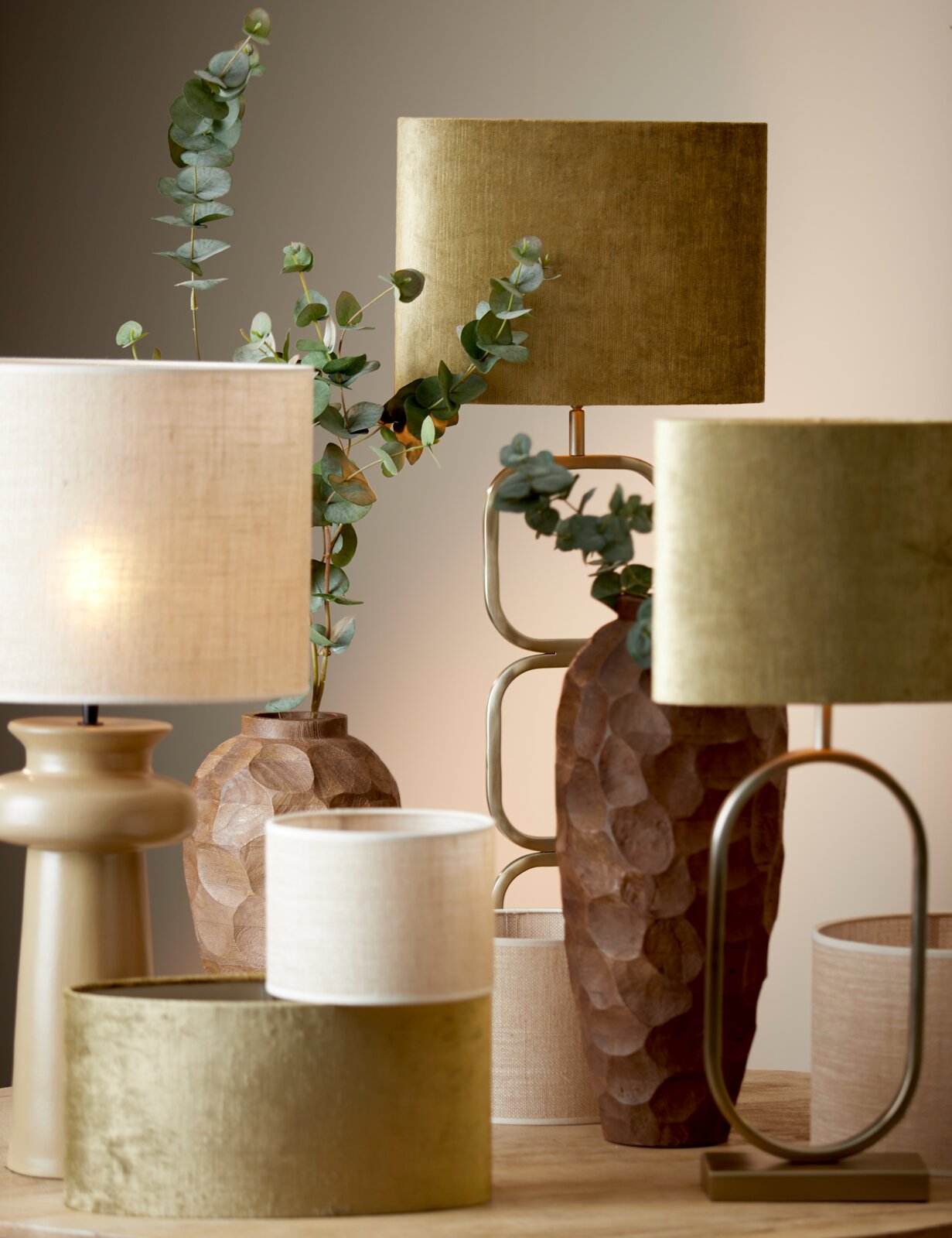 Verschillende lampenkappen in diverse kleuren en texturen, waaronder een gouden cilindervormige lampenkap op de voorgrond, omringd door decoratieve vazen en planten, gefotografeerd vanuit een zijdelingse hoek.