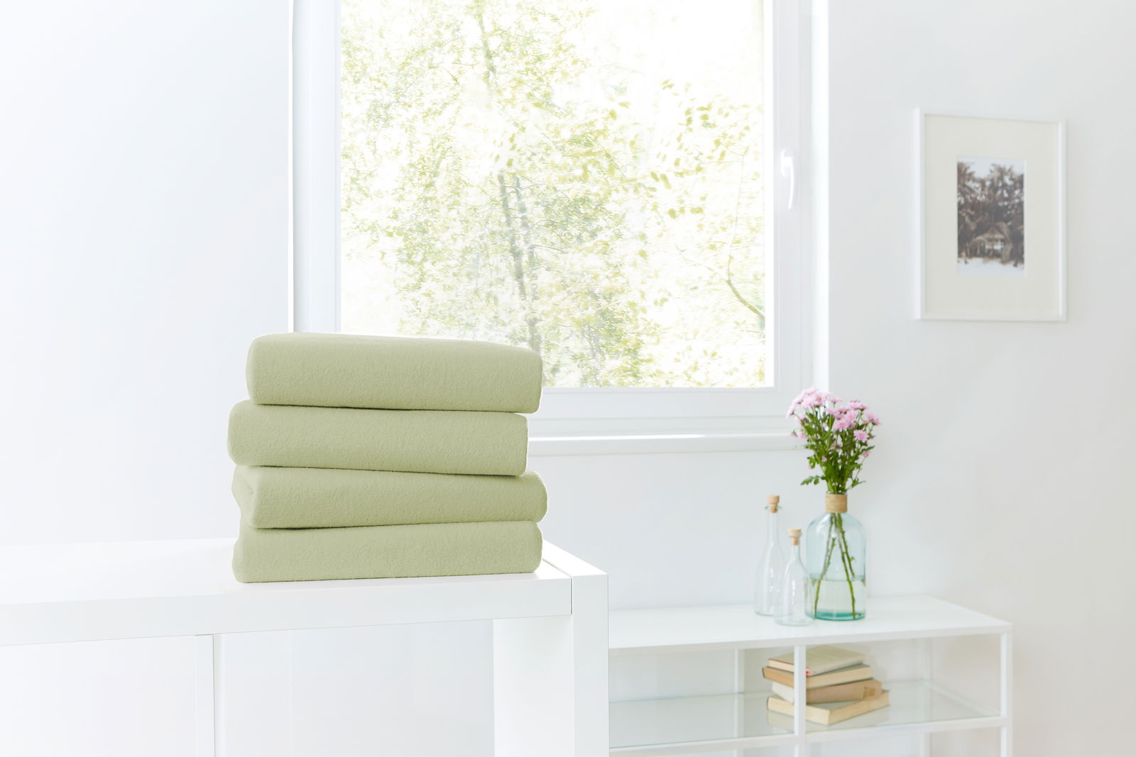 Novel Spannbetttuch TEDDY-FLAUSCH Ein Stapel von vier grünen Spannbetttüchern auf einem weißen Tisch, im Hintergrund ein Fenster mit Blick ins Grüne. Perspektive von vorne.