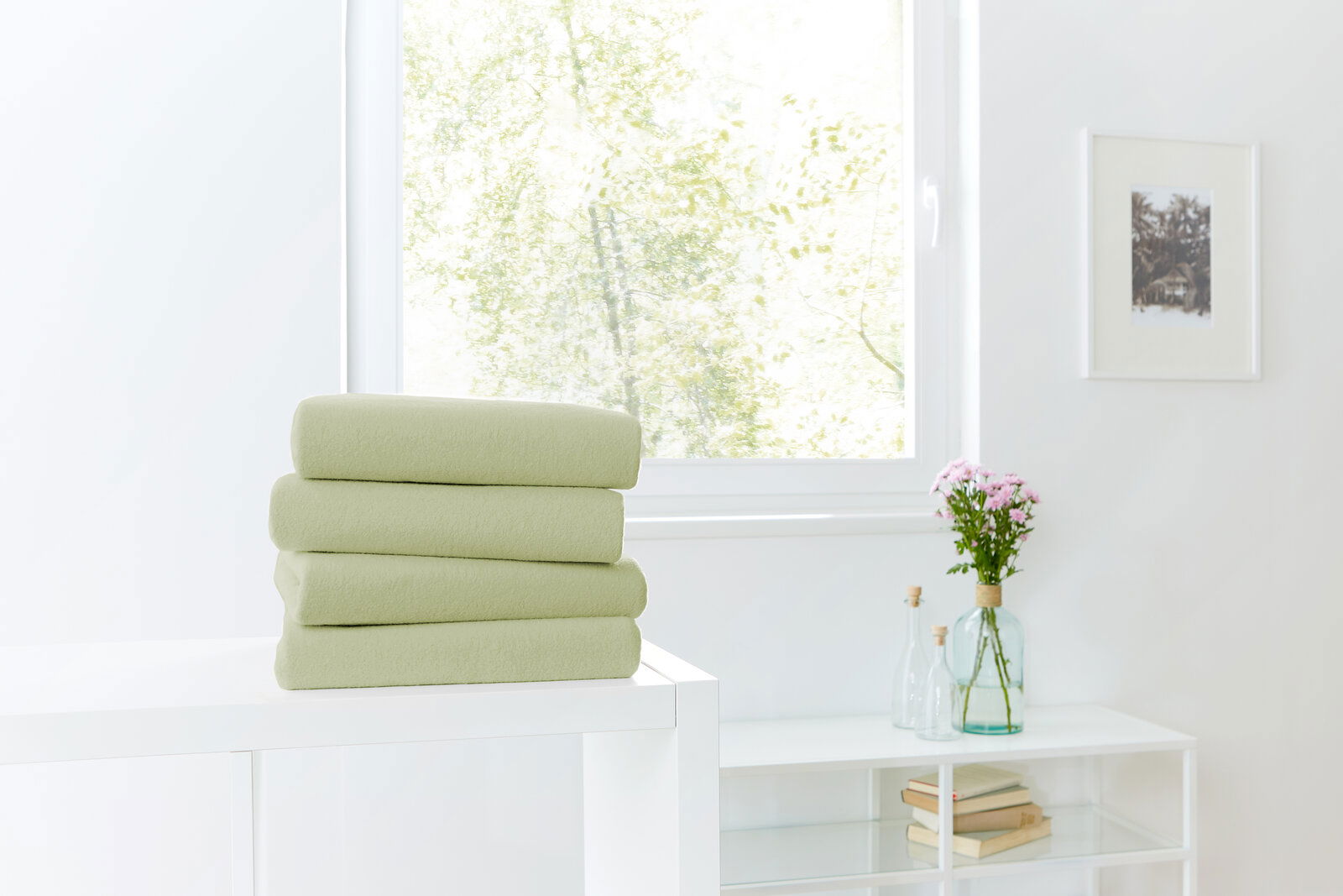 Novel Spannbetttuch TEDDY-FLAUSCH Ein Stapel von vier grünen Spannbetttüchern auf einem weißen Tisch, im Hintergrund ein Fenster mit Blick ins Grüne. Perspektive von vorne.