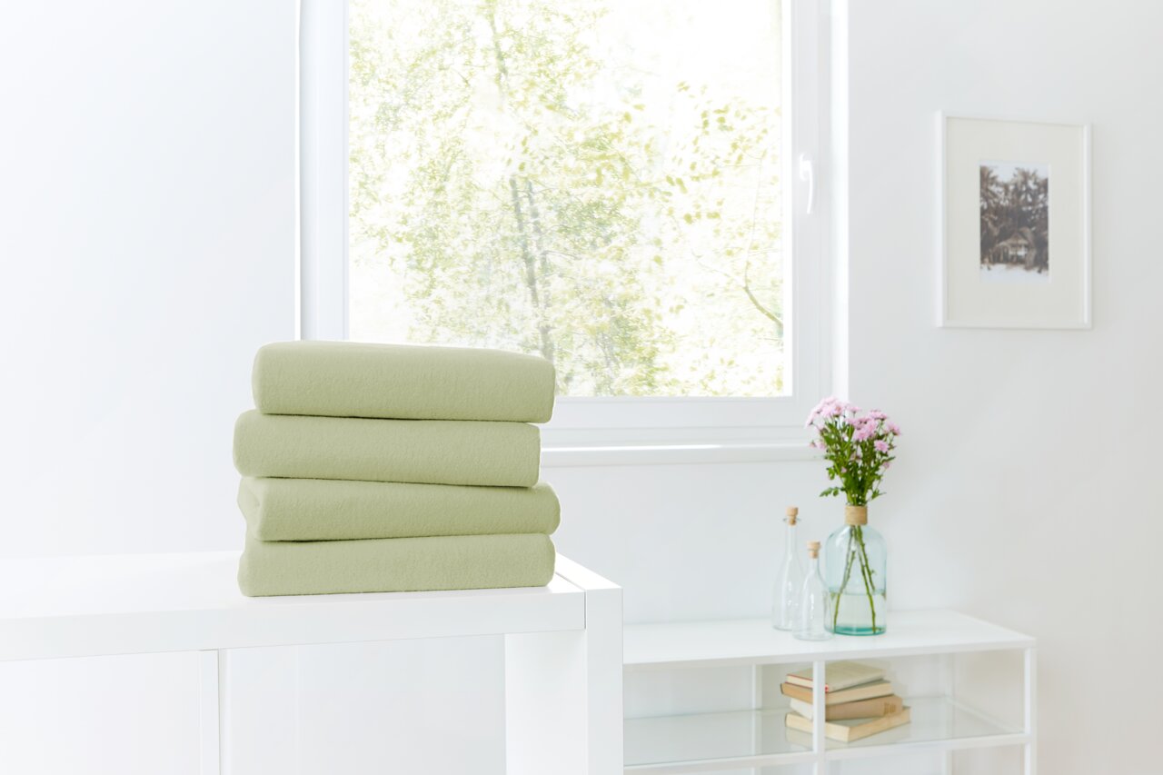 Ein Stapel von vier grünen Spannbetttüchern auf einem weißen Tisch, im Hintergrund ein Fenster mit Blick ins Grüne. Perspektive von vorne.