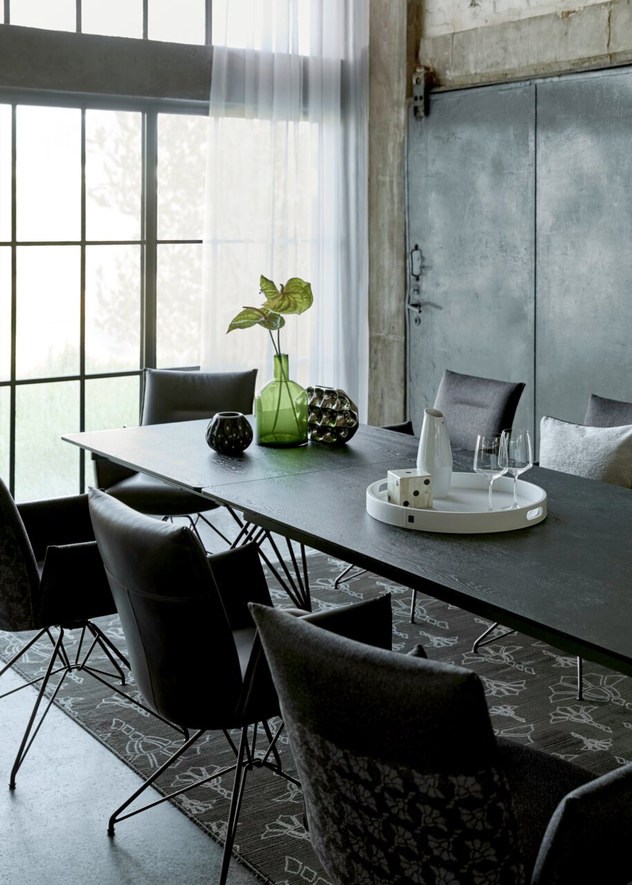 Schwarzer Esstisch im Industrial-Stil mit dunklen Polsterstühlen, dekoriert mit grüner Glasvase und Tablett mit Karaffe und Gläsern, aufgenommen aus schräger Seitenperspektive im hellen Raum mit großen Sprossenfenstern.