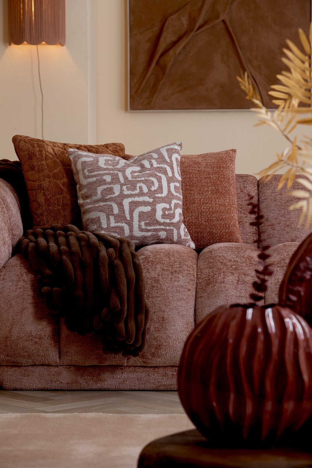 Vooraanzicht van een warme terracottakleurige bank met decoratieve sierkussens en een donkerbruine plaid in een sfeervol interieur.