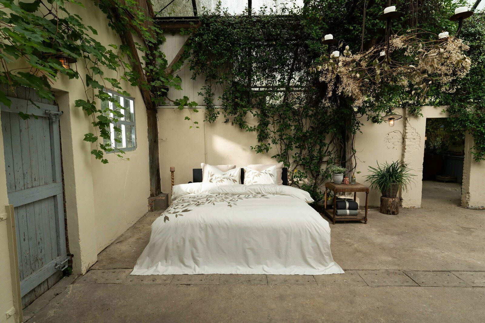 Bett mit weißer Bettwäsche und Blattmuster in einem rustikalen Raum mit Pflanzen und Holzmöbeln, aus frontaler Perspektive.