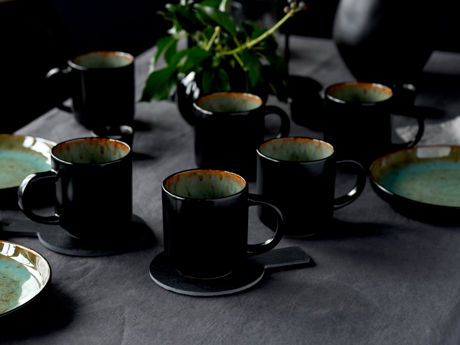 Schwarze Keramik-Kaffeetassen mit türkis glasierter Innenseite auf Untersetzern und gedecktem Tisch, schräg von vorn aus leicht erhöhter Perspektive fotografiert