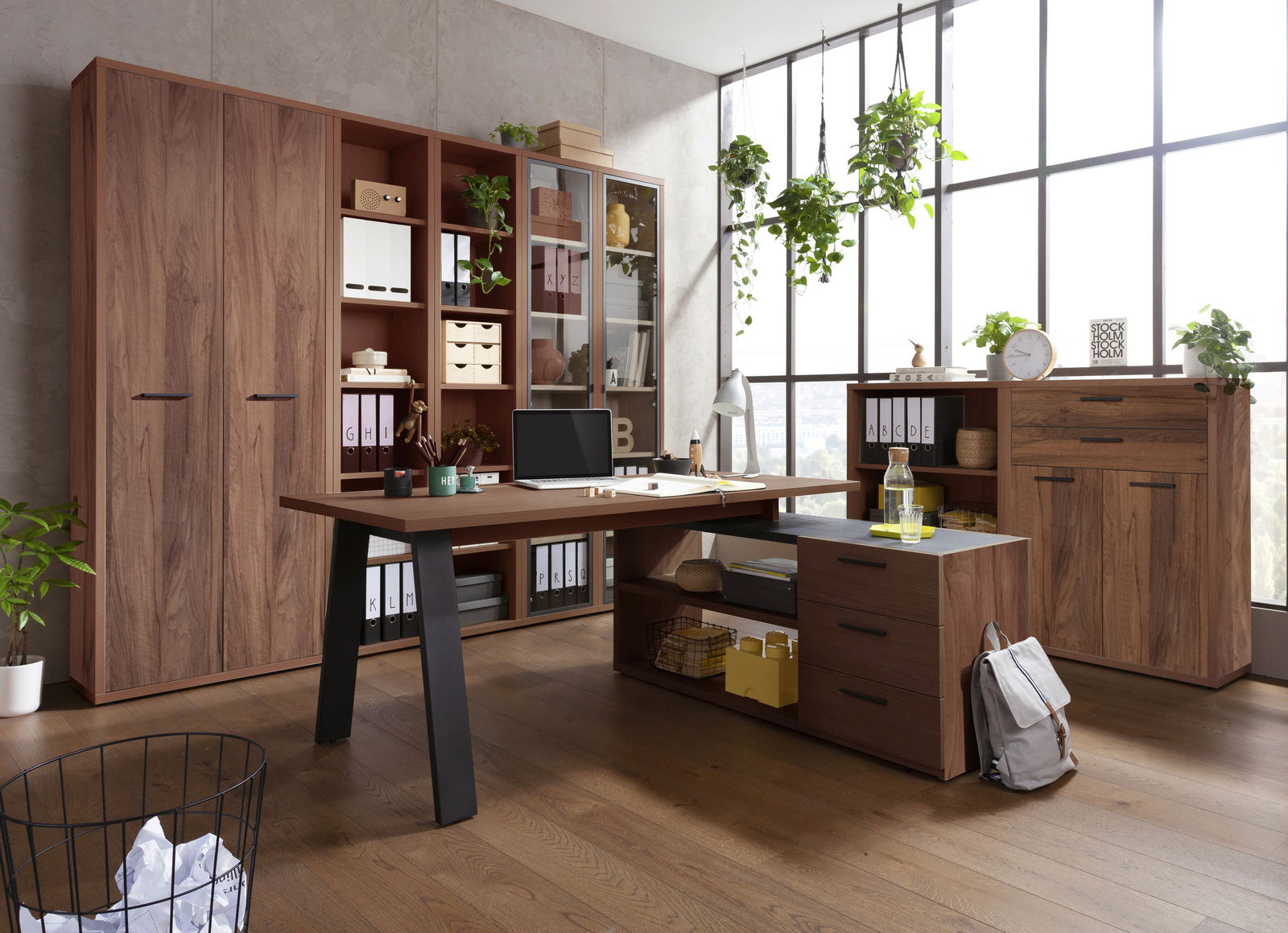 Moderne Büromöbel in einem hellen Raum mit großen Fenstern, bestehend aus einem Eckschreibtisch, Regalen und Schränken in Holzoptik, mit Pflanzen und Dekorationen. Perspektive von vorne.