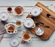 Vogelperspektive auf ein Set von vier elfenbeinfarbenen Mini-Cocottes, gefüllt mit Dessert und umgeben von Kaffee, Zuckerwürfeln und Löffeln auf einem Holztablett.