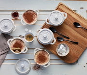 Vogelperspektive auf ein Set von vier elfenbeinfarbenen Mini-Cocottes, gefüllt mit Dessert und umgeben von Kaffee, Zuckerwürfeln und Löffeln auf einem Holztablett.