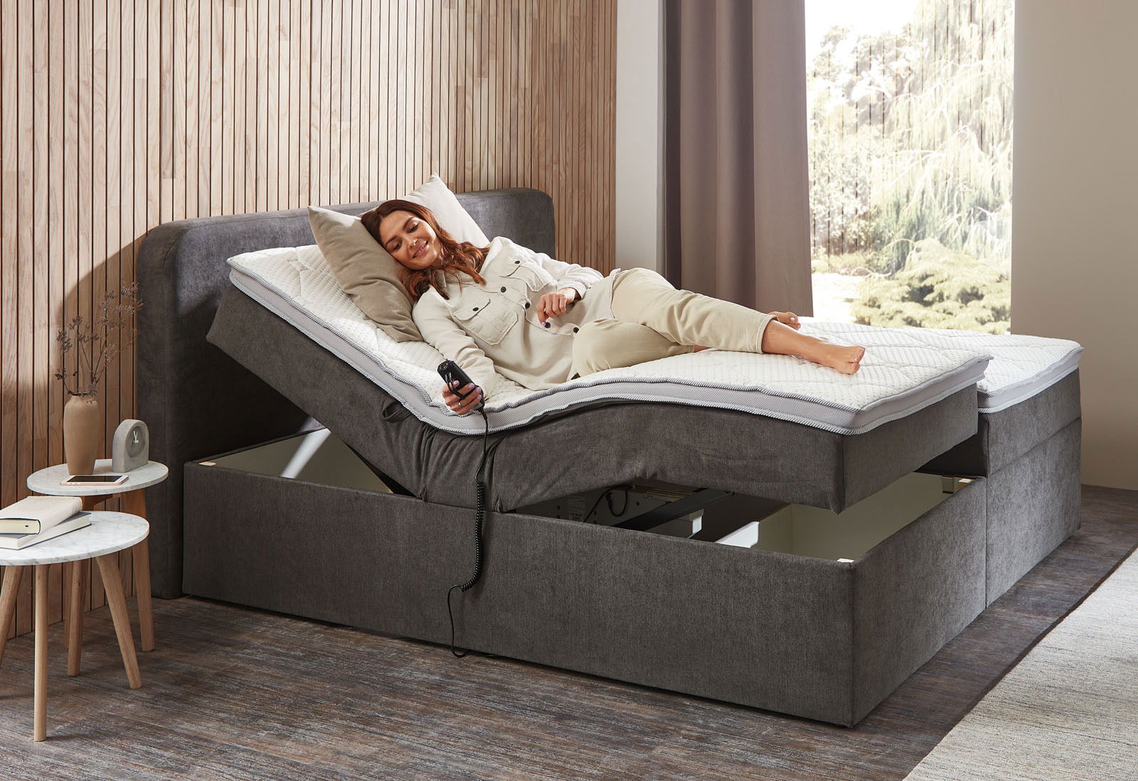 Boxbett mit Motor in seitlicher Perspektive, eine Person liegt entspannt darauf und hält eine Fernbedienung.