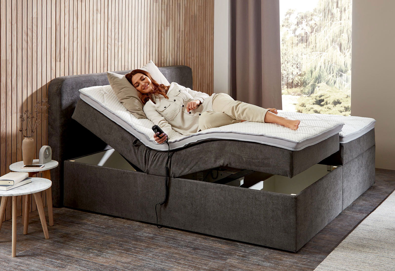 Boxbett mit Motor in seitlicher Perspektive, eine Person liegt entspannt darauf und hält eine Fernbedienung.