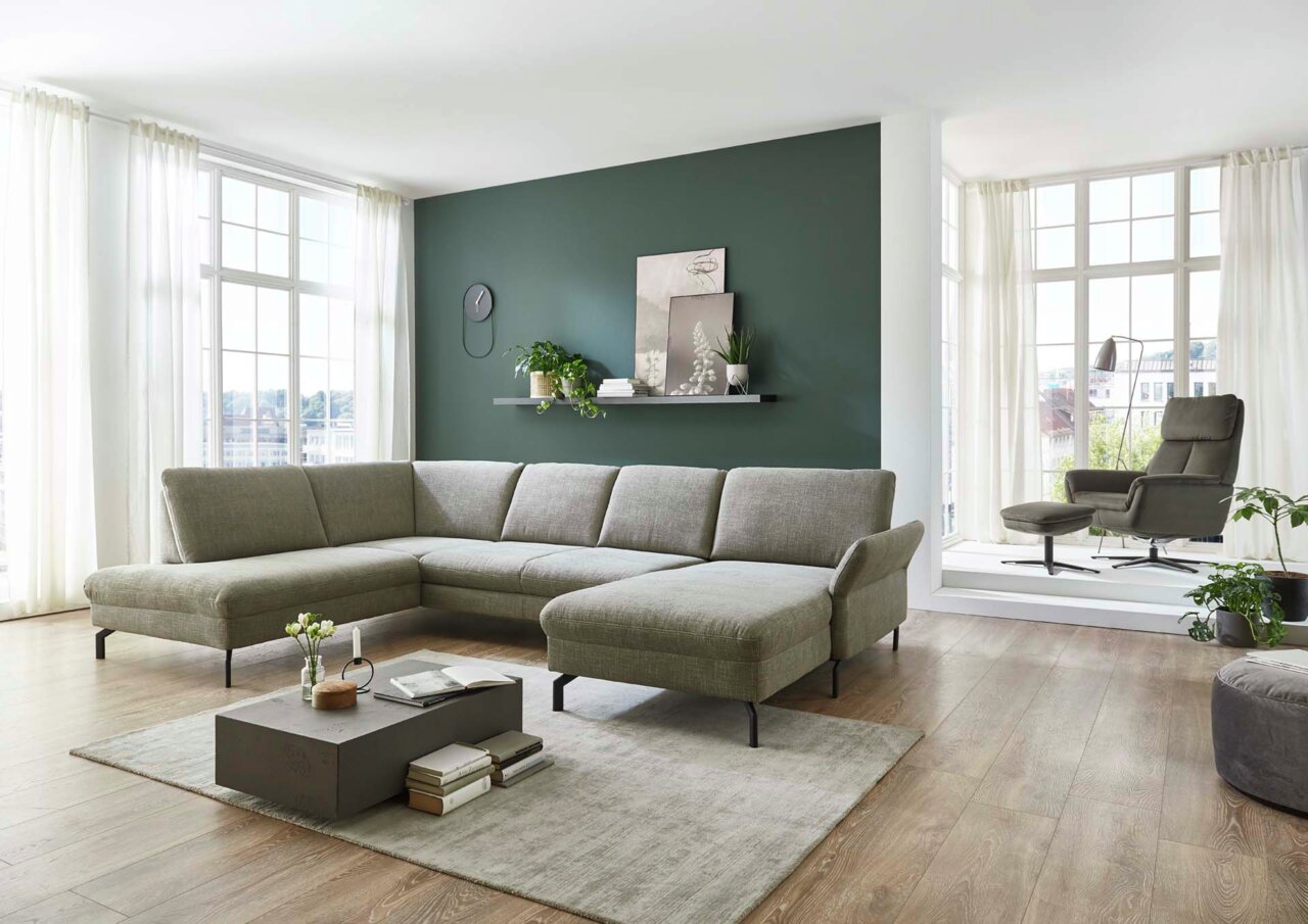 Moderne Polsterrundecke in einem hellen Wohnzimmer mit großen Fenstern, grauer Wand und stilvollem Dekor. Perspektive von vorne links.