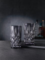 Zwei elegante Longdrinkgläser aus Kristallglas mit facettiertem Design, frontal auf einem dunklen Hintergrund präsentiert.