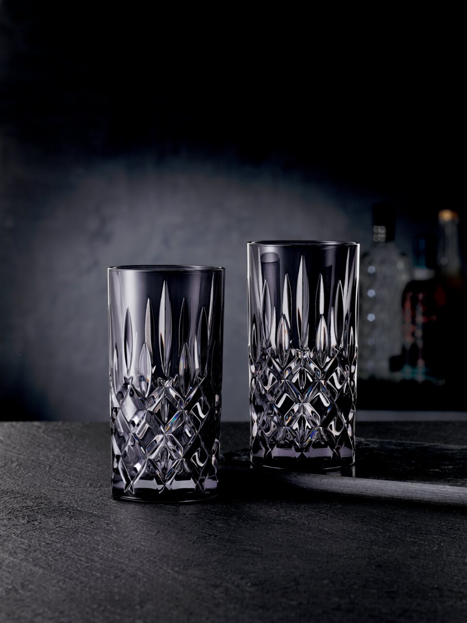 Twee elegante longdrinkglazen van kristalglas met een gefacetteerd ontwerp, frontaal afgebeeld tegen een donkere achtergrond.