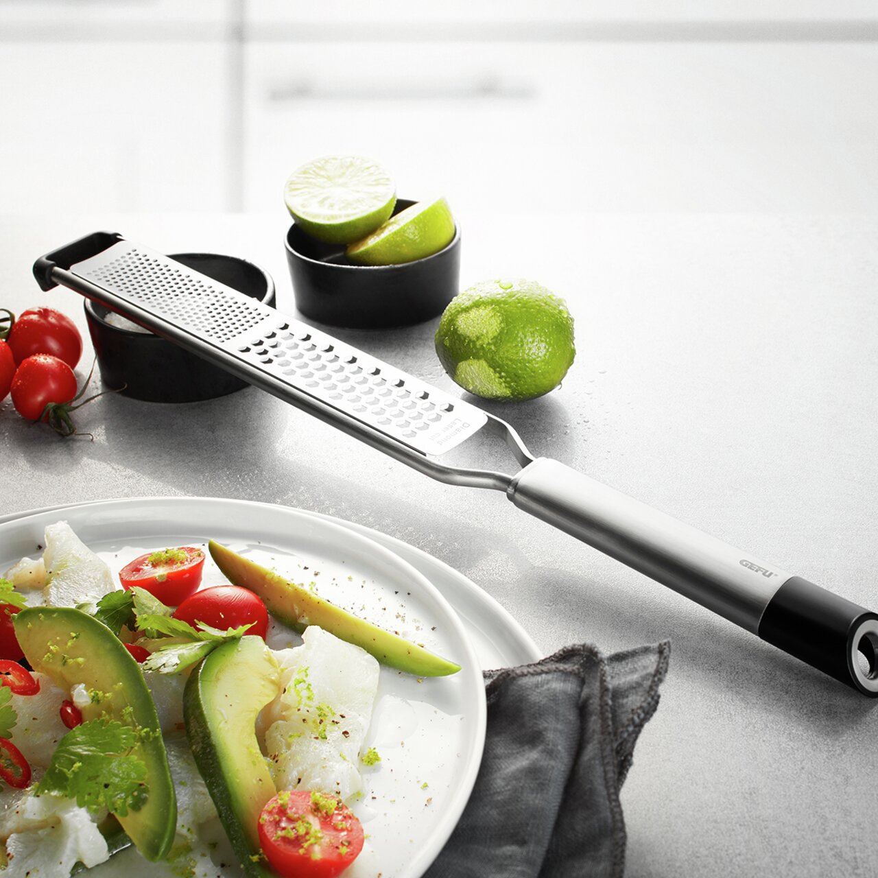 Fein- und Grobreibe PRIMELINE aus Edelstahl, seitliche Perspektive, auf einer Küchenarbeitsplatte mit Limetten und Tomaten im Hintergrund.