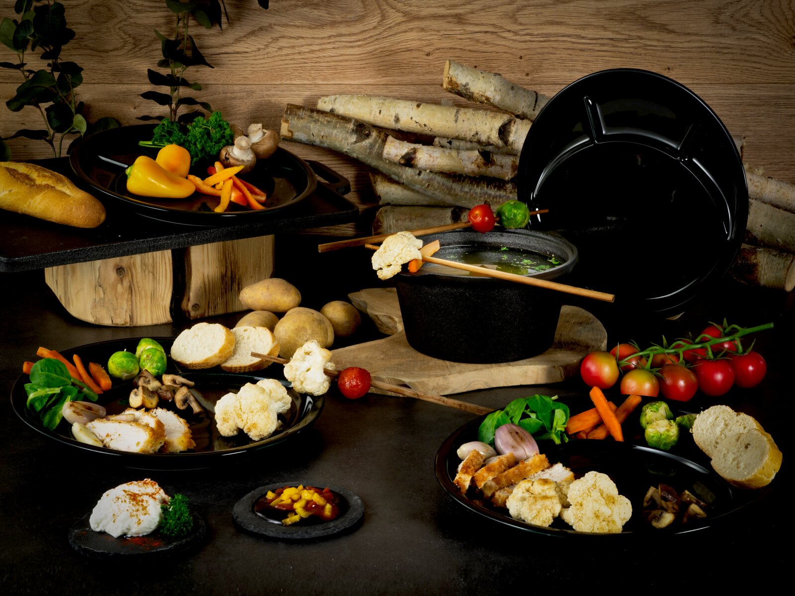 Schwarzes Fondue-Set mit Topf, Deckel und Tellern, angerichtet mit Gemüse, Brot und Spießen auf rustikalem Holzhintergrund, perspektivisch in leichter Seitenansicht.