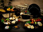 Schwarzes Fondue-Set mit Topf, Deckel und Tellern, angerichtet mit Gemüse, Brot und Spießen auf rustikalem Holzhintergrund, perspektivisch in leichter Seitenansicht.