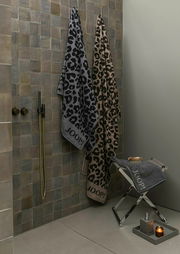 Zwei Saunatücher mit Leopardenmuster in Grau und Braun hängen an einer Wand, daneben ein weiteres Tuch auf einem Hocker. Perspektive: Badezimmeransicht.