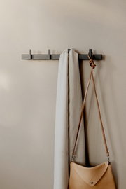 Frontale Ansicht einer minimalistischen schwarzen Wandgarderobe mit mehreren Haken, daran ein helles Tuch und eine beige Leder-Umhängetasche vor neutraler Wand