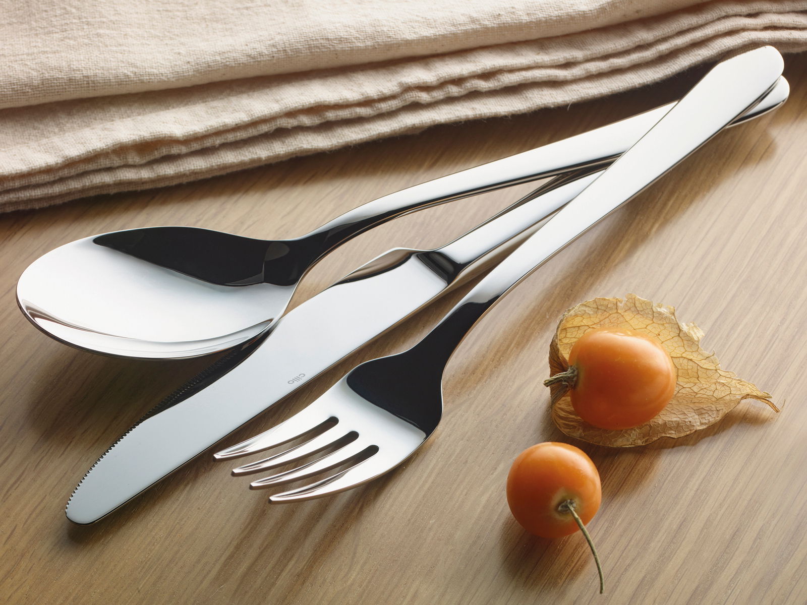 Elegantes Besteck-Set bestehend aus Löffel, Messer und Gabel in Nahaufnahme auf einem Holztisch, begleitet von Physalis-Früchten und einem beigen Stofftuch.