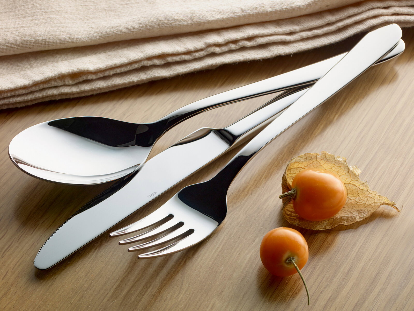 Elegantes Besteck-Set bestehend aus Löffel, Messer und Gabel in Nahaufnahme auf einem Holztisch, begleitet von Physalis-Früchten und einem beigen Stofftuch.