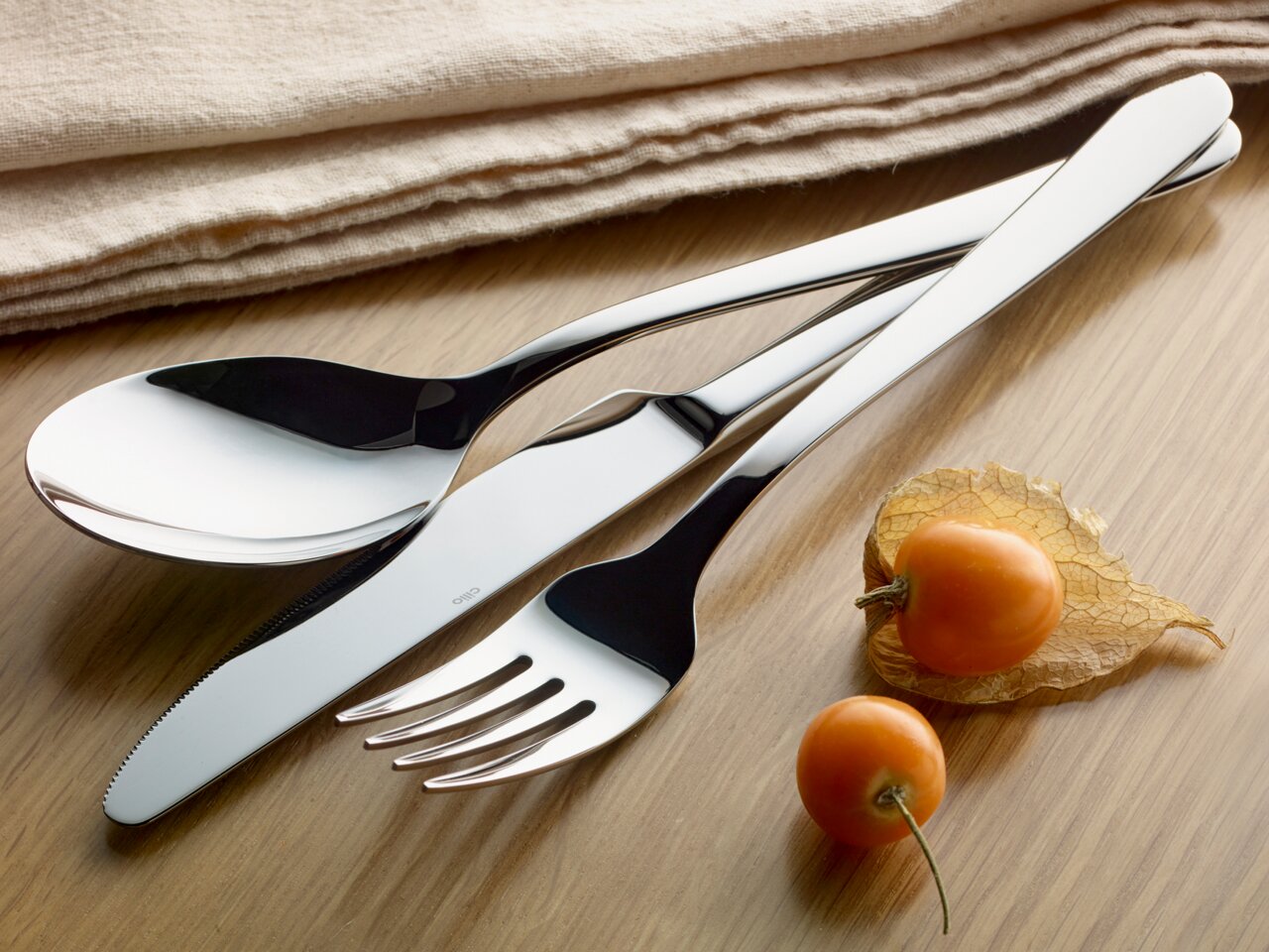 Cilio Besteck-Set 30-tlg. ROMA Elegantes Besteck-Set bestehend aus Löffel, Messer und Gabel in Nahaufnahme auf einem Holztisch, begleitet von Physalis-Früchten und einem beigen Stofftuch.