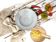 Hellblaue, gesprenkelte Keramikschüssel mit braunem Rand auf rustikalem, weiß verwittertem Holztisch, Draufsicht; daneben Leinentücher, Löffel, Chili, Tomaten und Tagliatelle auf Holzbrett.