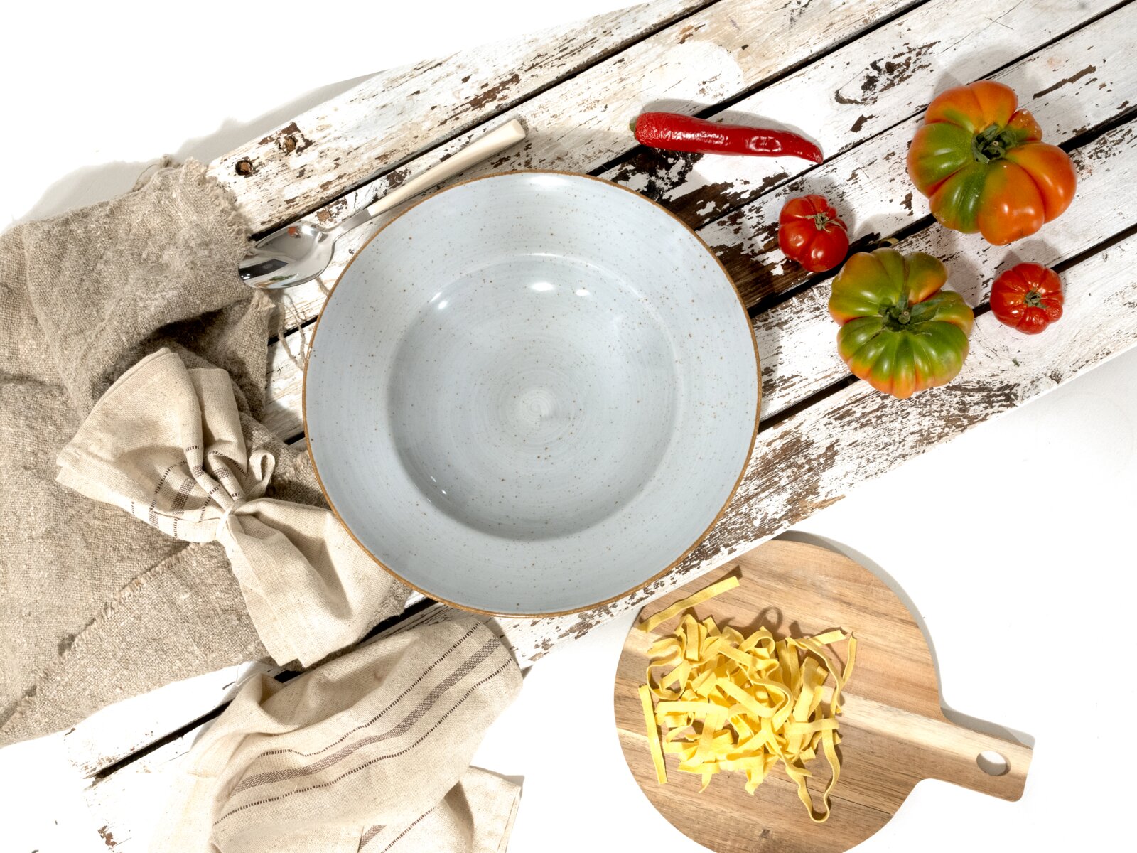 Hellblaue, gesprenkelte Keramikschüssel mit braunem Rand auf rustikalem, weiß verwittertem Holztisch, Draufsicht; daneben Leinentücher, Löffel, Chili, Tomaten und Tagliatelle auf Holzbrett.